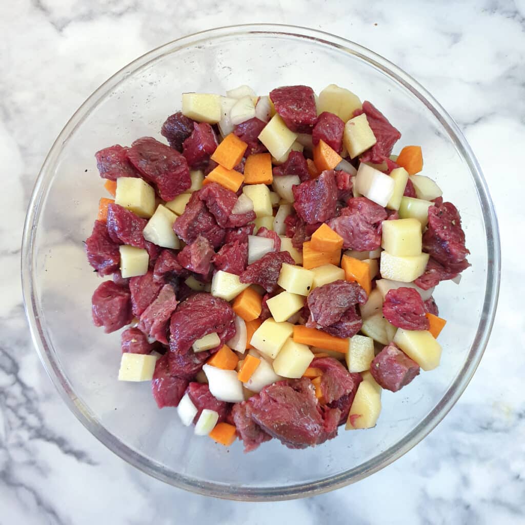A glass bowl containing cubed beef, potatoes, carrots and onions.