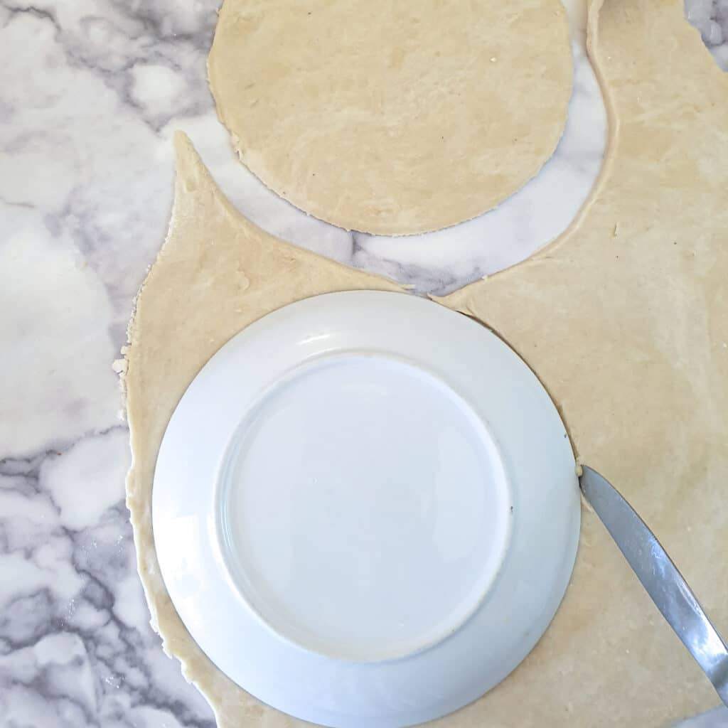 Dough rolled out thinly and being cut around a side plate to form a large circle.