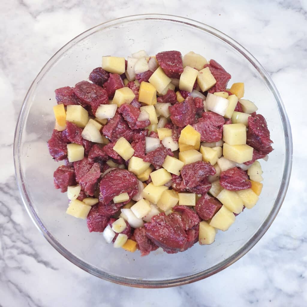 A glass bowl containing cubed beef, potatoes, carrots and onions.