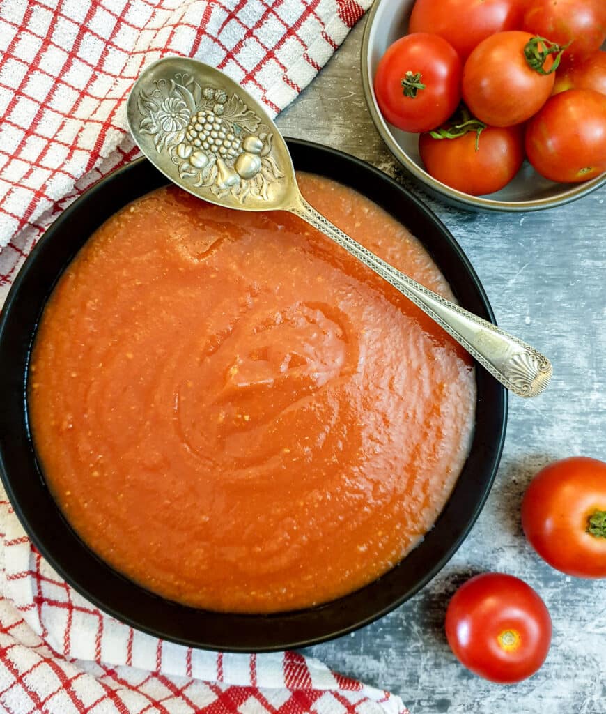 A black bowl of homemade tomato passata alongside a bowl of fresh tomatoes and a red-checked tea towel.