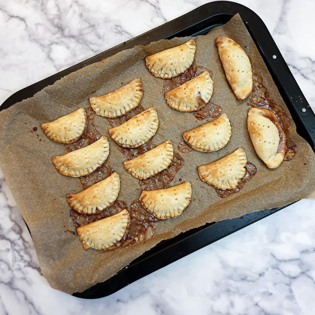 Baked jam turnovers on a baking sheet with a small amount of jam spilling out.