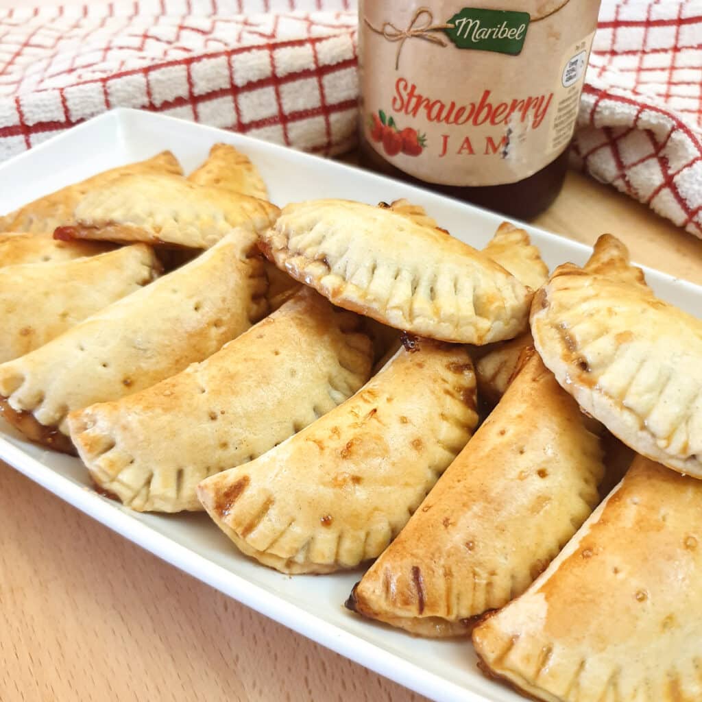 A long white serving plate filled with jam turnovers in front of a jar of strawberry jam.