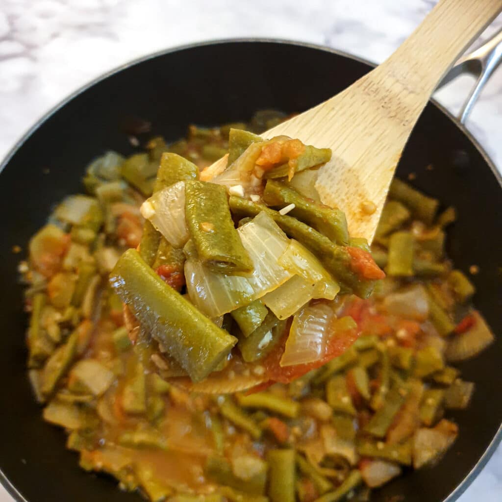 Green beans on a wooden spoon held over a frying pan of Middle Eastern-style runner beans.