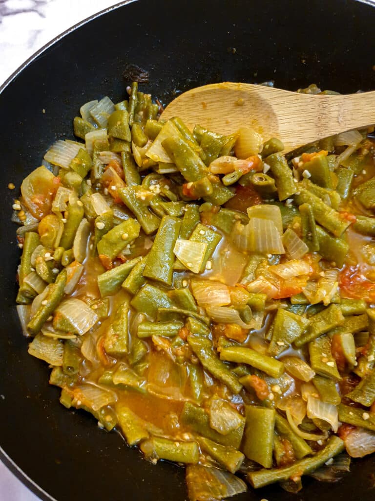 A wooden spoon stirring a pan of runner beans, tomatoes and onions.