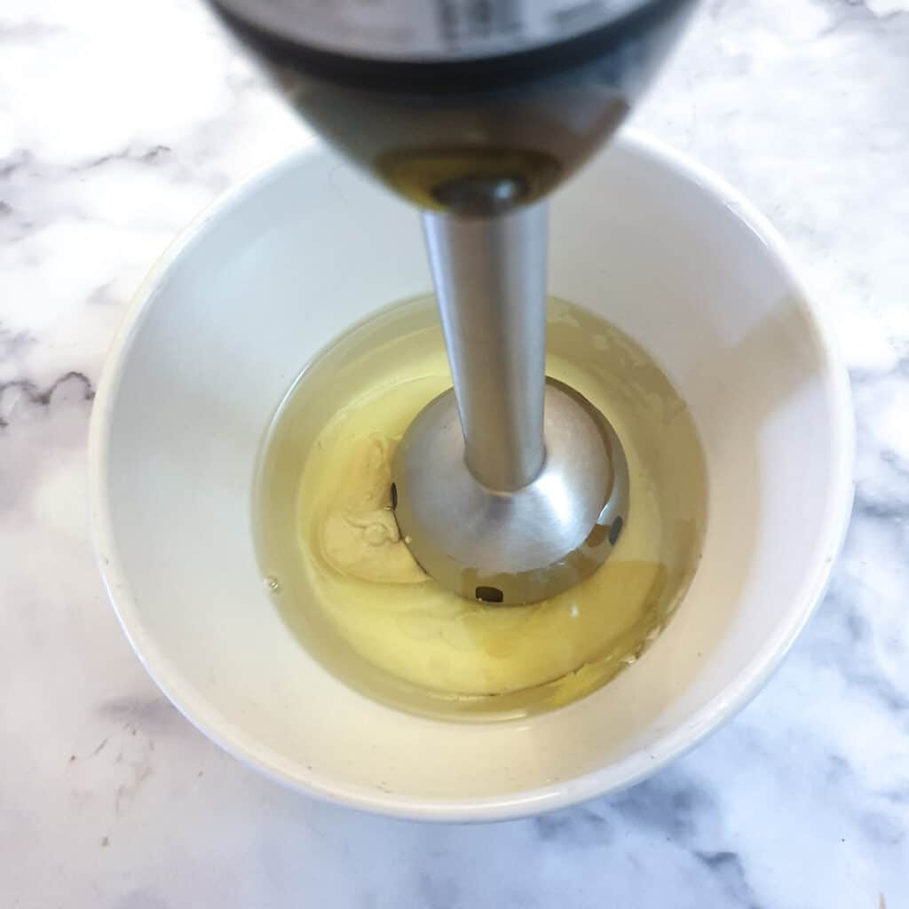 A stick blender covering an egg in a dish of oil.