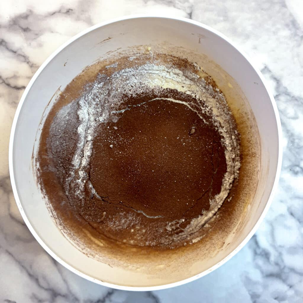 Milk, flour and cocoa powder sieved on top of the egg and milk mixture in a white bowl.