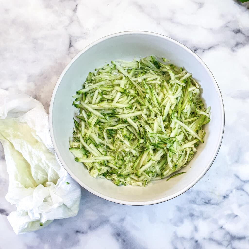 A dish of grated courgettes next to a paper towel that was used to press out the moisture.