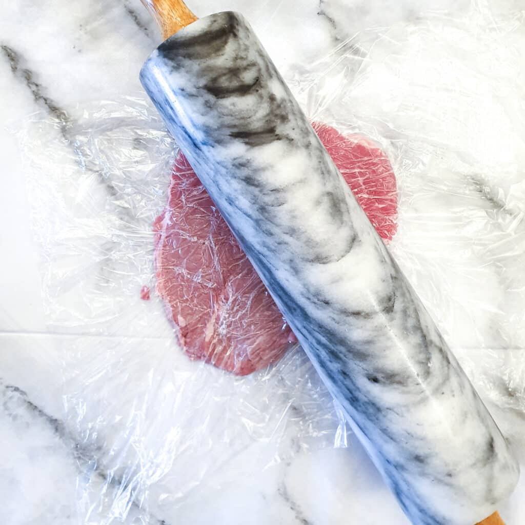 A beef schnitzel being flattened with a rolling pin between layers of plastic wrap.