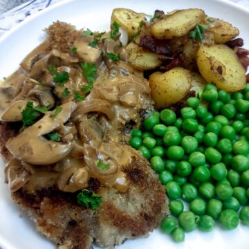 A crispy crumbed beef schnitzel on a plate with potatoes, peas and a mushroom sauce.