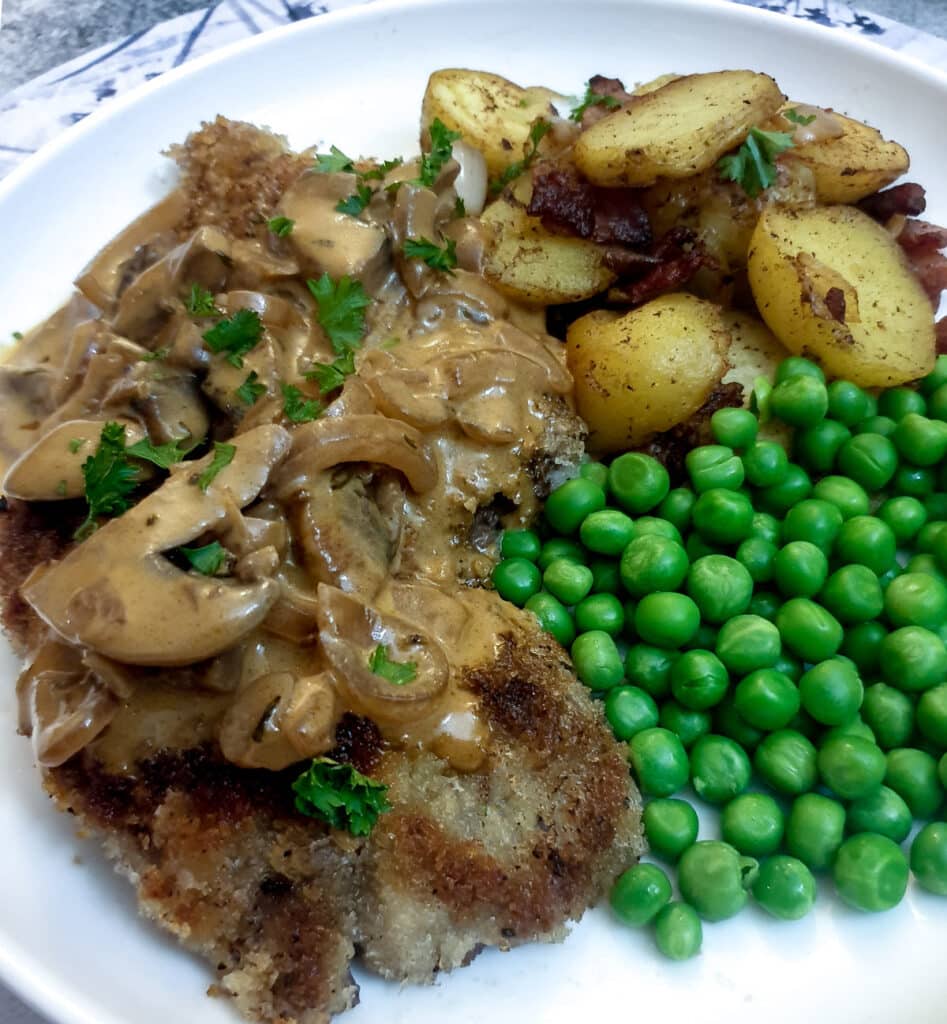 A crispy crumbed beef schnitzel on a plate with potatoes, peas and a mushroom sauce.