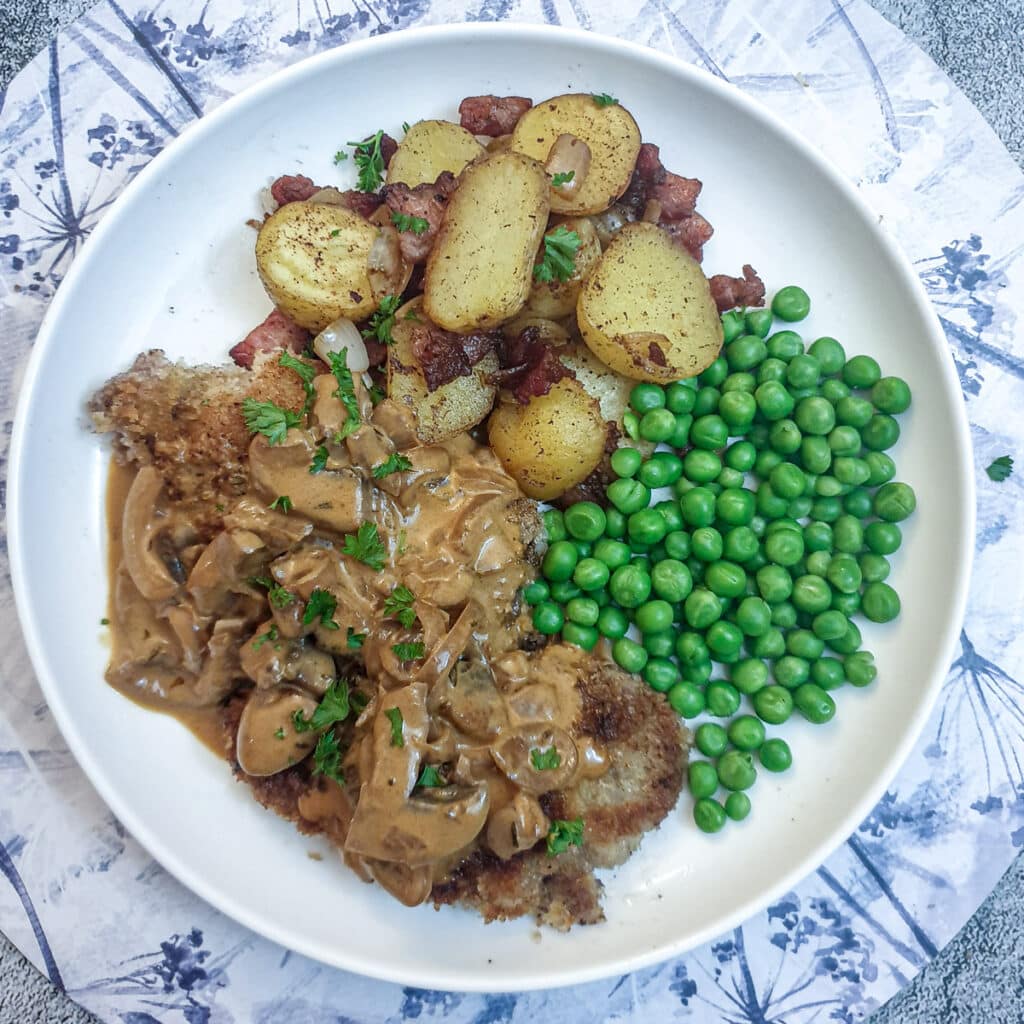 Overhead shot of a crispy beef schnitzel on a white plate with peas, potatoes and mushroom sauce.