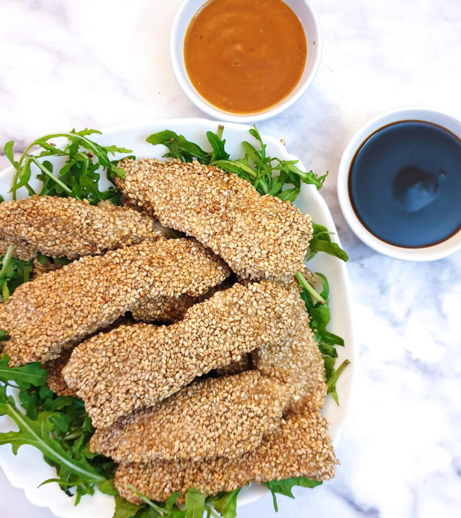 A pile of sesame-coated chicken strips on a bed of rocket next to 2 dipping sauces.
