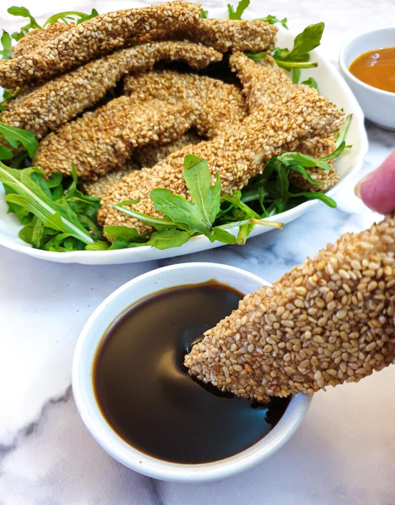 A piece of sesame-coated chicken being dipped into a bowl of dipping sauce.