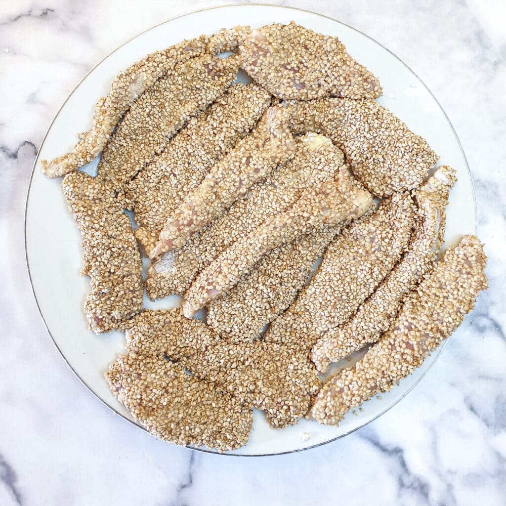 A plate of chicken strips which have been coated in egg and sesame seeds.