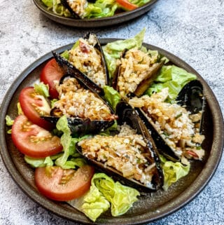 5 stuffed mussels on a brown plate garnished with shredded lettuce and sliced tomatoes.