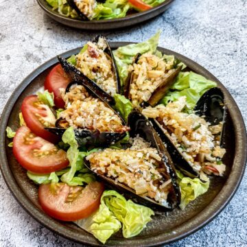 5 stuffed mussels on a brown plate garnished with shredded lettuce and sliced tomatoes.