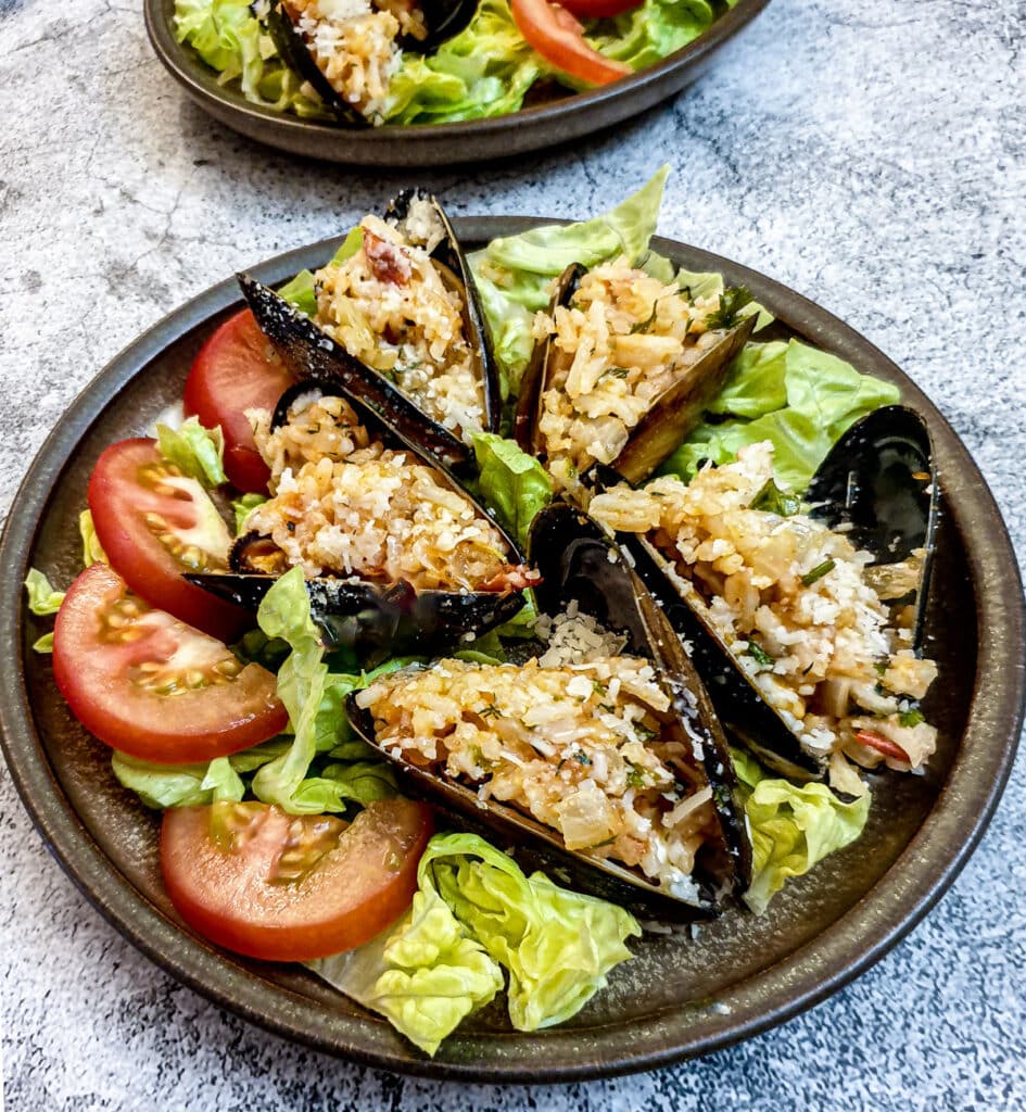 5 stuffed mussels on a brown plate garnished with shredded lettuce and sliced tomatoes.