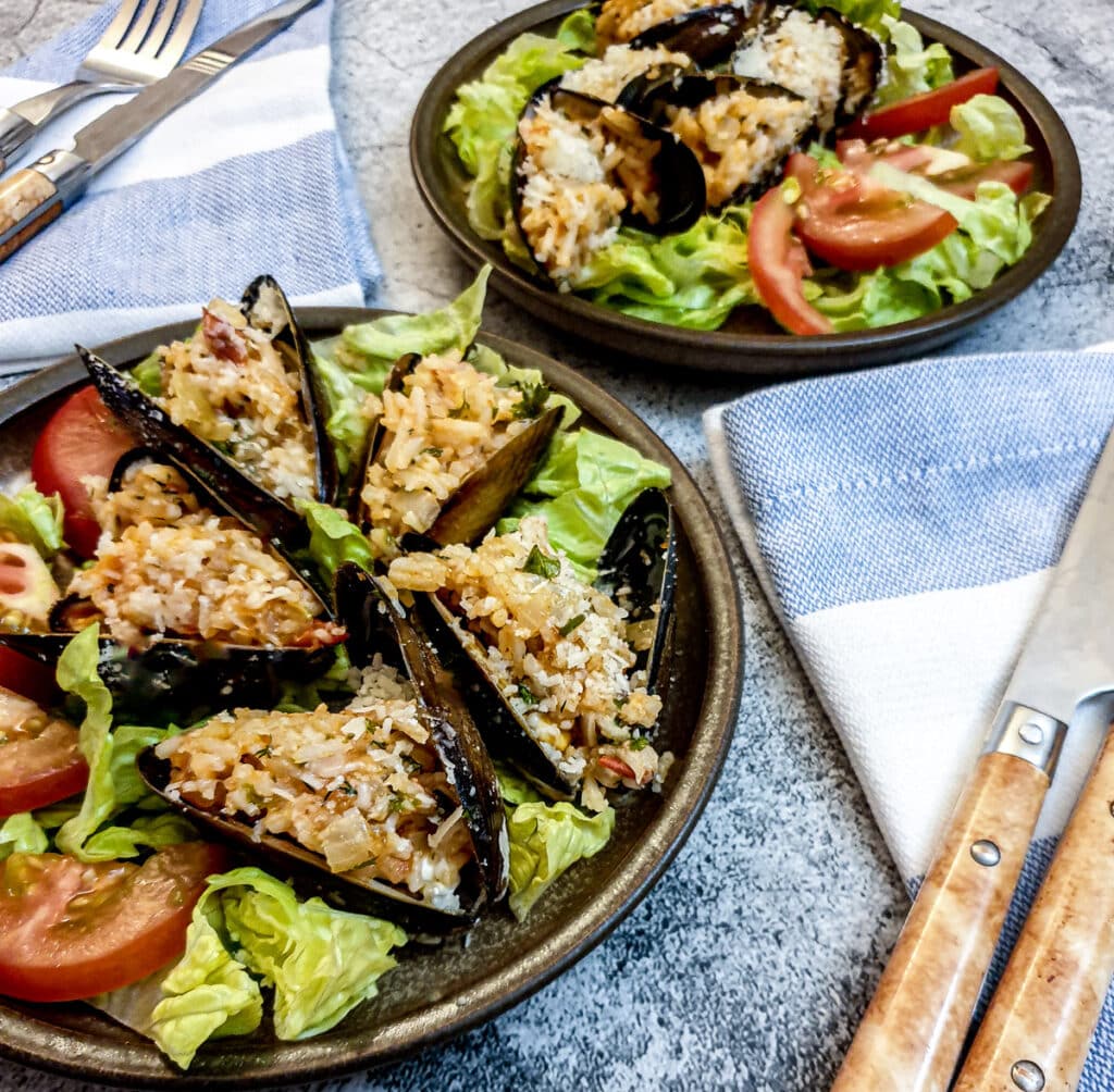 Stuffed mussels on a brown plate, with tomato and shredded lettuce, on a table.