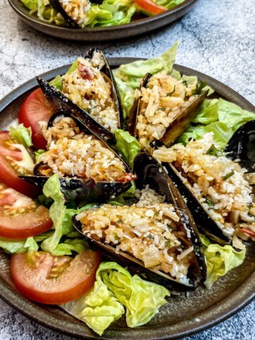 5 stuffed mussels on a brown plate garnished with shredded lettuce and sliced tomatoes.