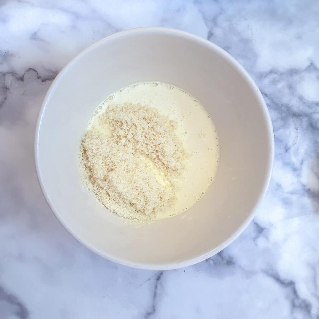 A small white bowl containing beaten egg, cream and milk with grated parmesan cheese on top.