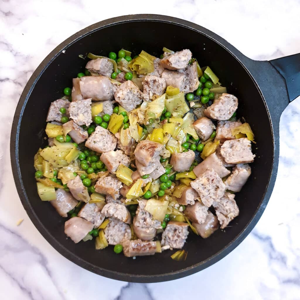 Chopped sausages, leeks and peas in a frying pan.