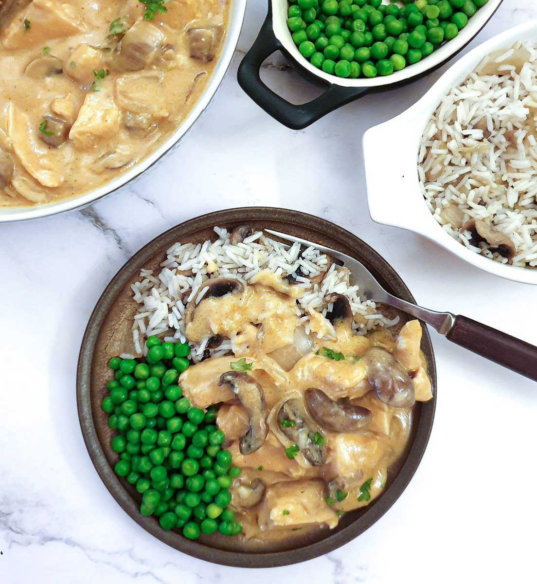 A brown dinner plate on a table, containing a serving of chicken Diane on a bed of rice, with peas on the side.  The serving dishes are in the background.