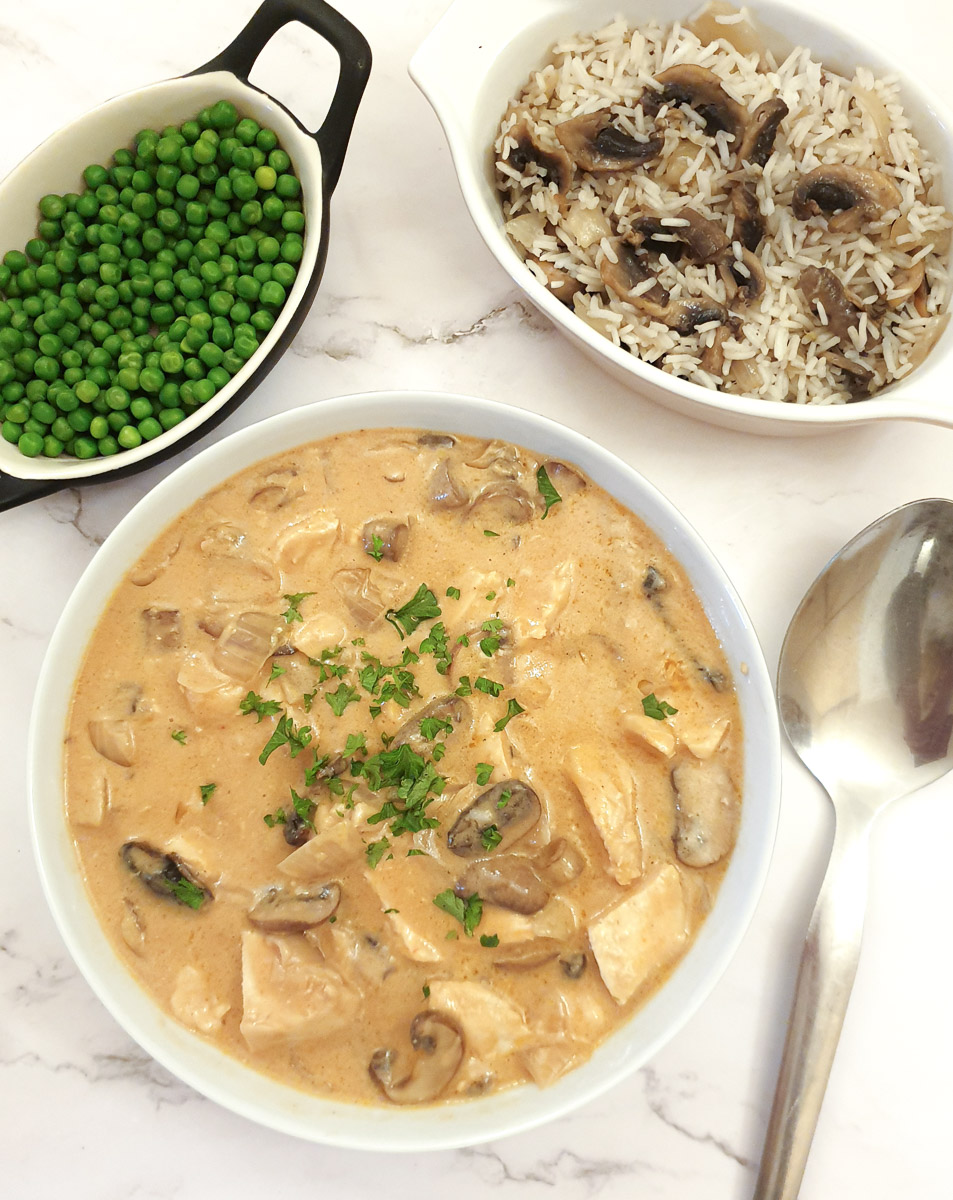 Chicken Diane in a white serving bowl on a table next to a serving spoon. There is also a dish of peas and a small dish of mushroom rice on the table.