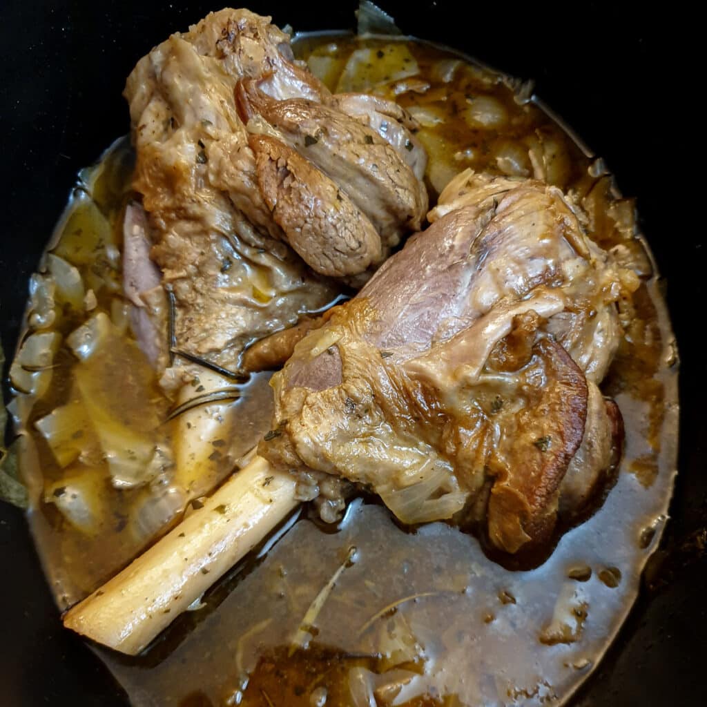 2 cooked lamb shanks in a slow cooker showing how the meat has pulled away from the bone.