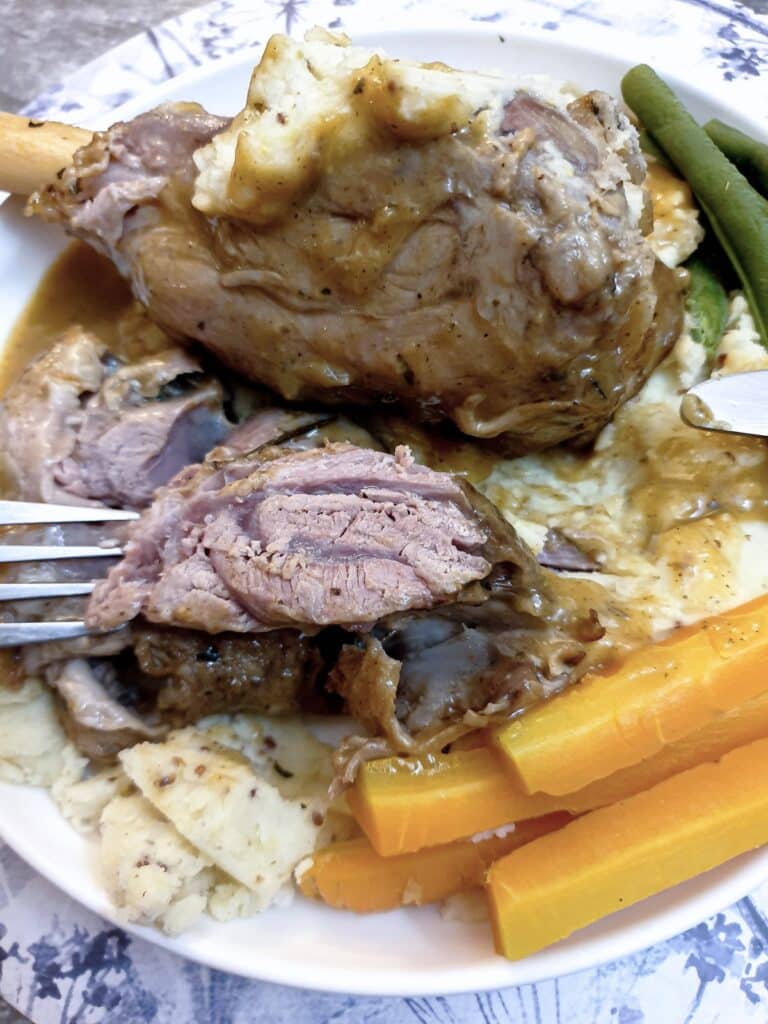 A piece of minted lamb shank on a fork held over a plate of lamb shank, potatoes and vegetables.