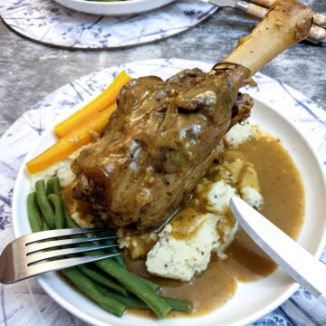 A minted lamb shank on a bed of potatoes with vegetables on the side.