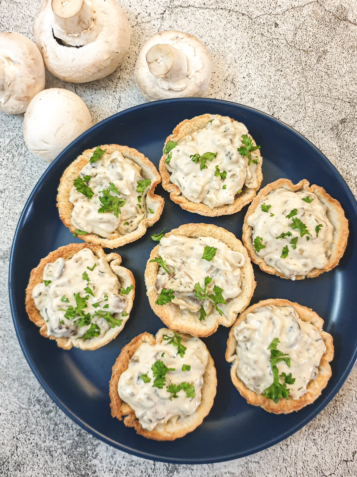 Overhead shot of 7 mushroom breadcups on a plate next to fresh mushrooms.
