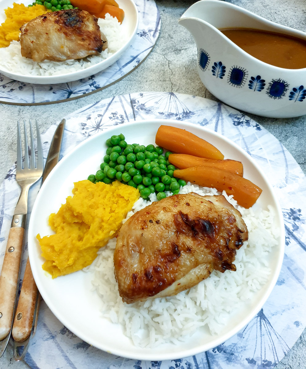 A white dinner plate with a chicken thigh on a pile of rice, with carrots, peas and pumpkin vegetables.  There is a gravy boat of sauce in the background.