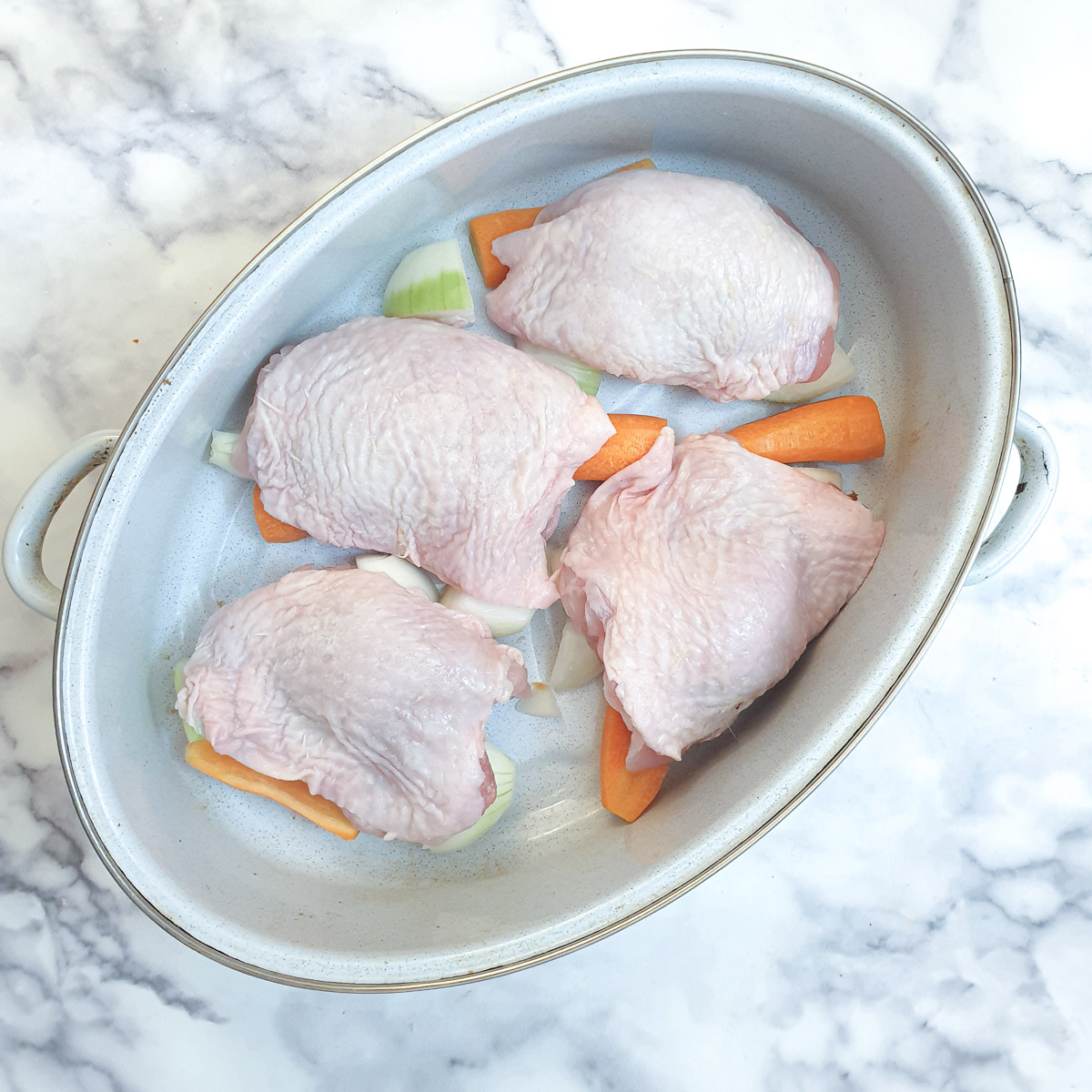 4 chicken thighs on top of chopped carrots and onions in a light blue roasting dish.