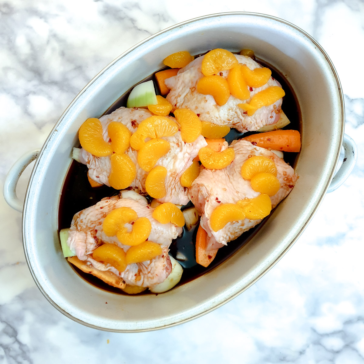 4 raw chicken thighs covered with orange segments and gravy in a light blue roasting pan.