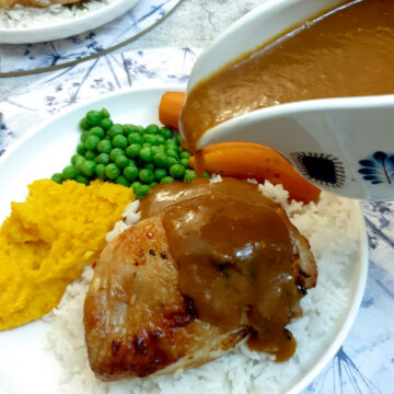 A sauce boat of orange sauce being poured over a plate of chicken thighs and vegetables.