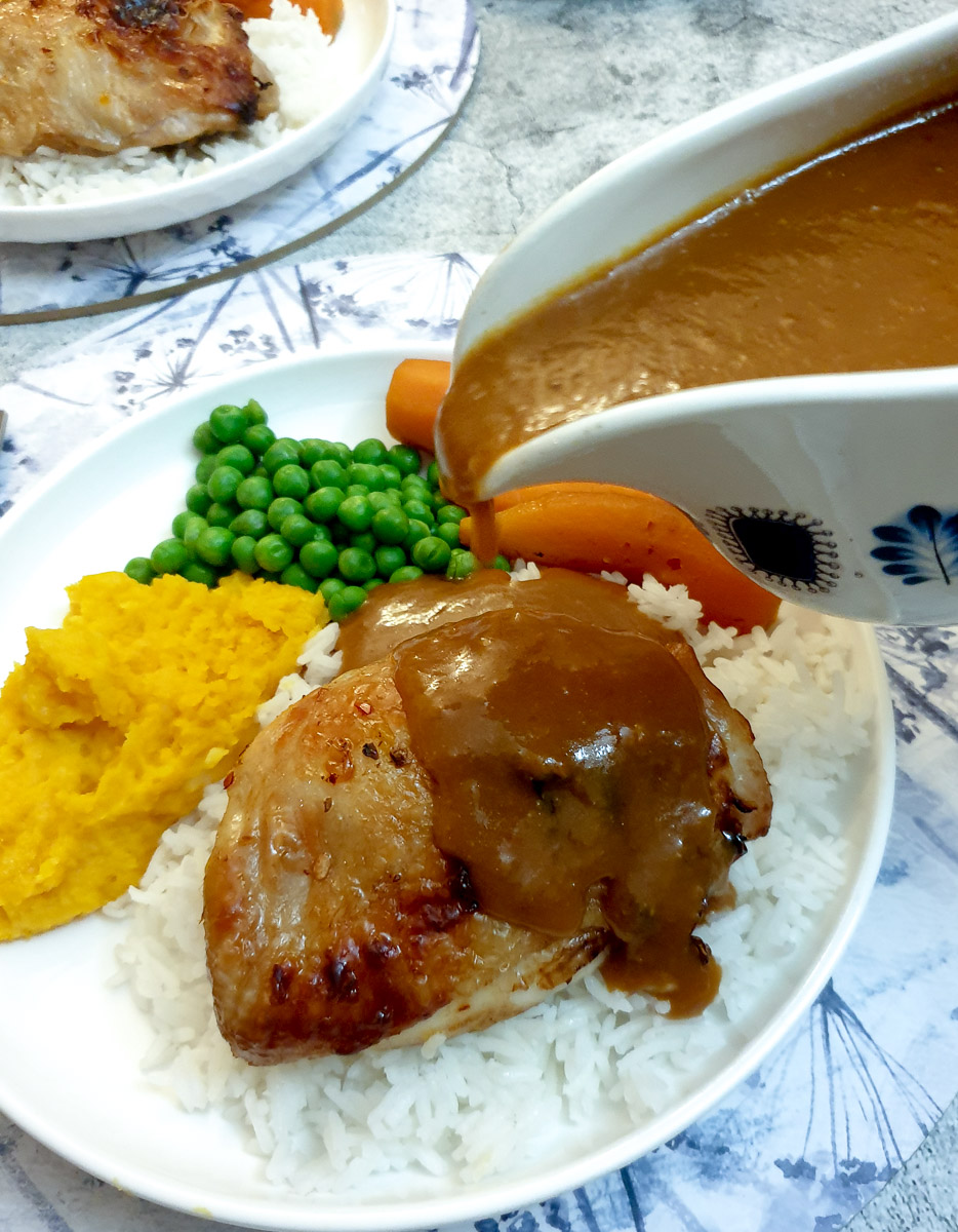 A sauce boat of orange sauce being poured over a plate of chicken thighs and vegetables.