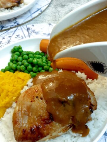 A sauce boat of orange sauce being poured over a plate of chicken thighs and vegetables.