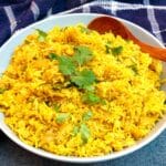 A white serving dish holding yellow pilau rice and a wooden serving spoon.