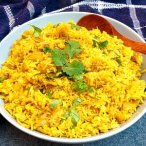 A white serving dish holding yellow pilau rice and a wooden serving spoon.