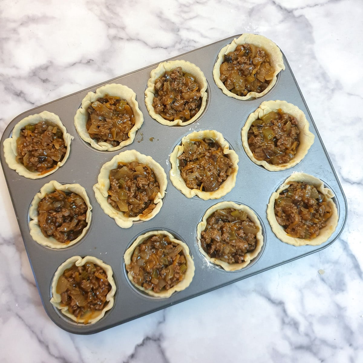 12 beef and onion pies in a muffin tray without pastry lids.