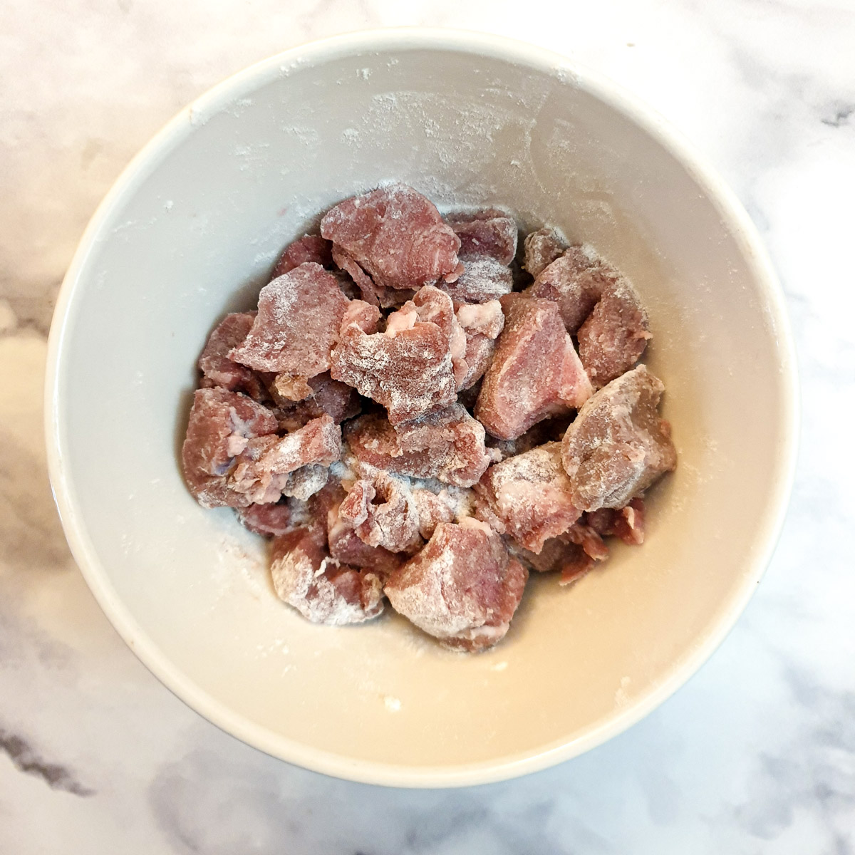 Cubes of lamb coated in flour in a small white mixing bowl.