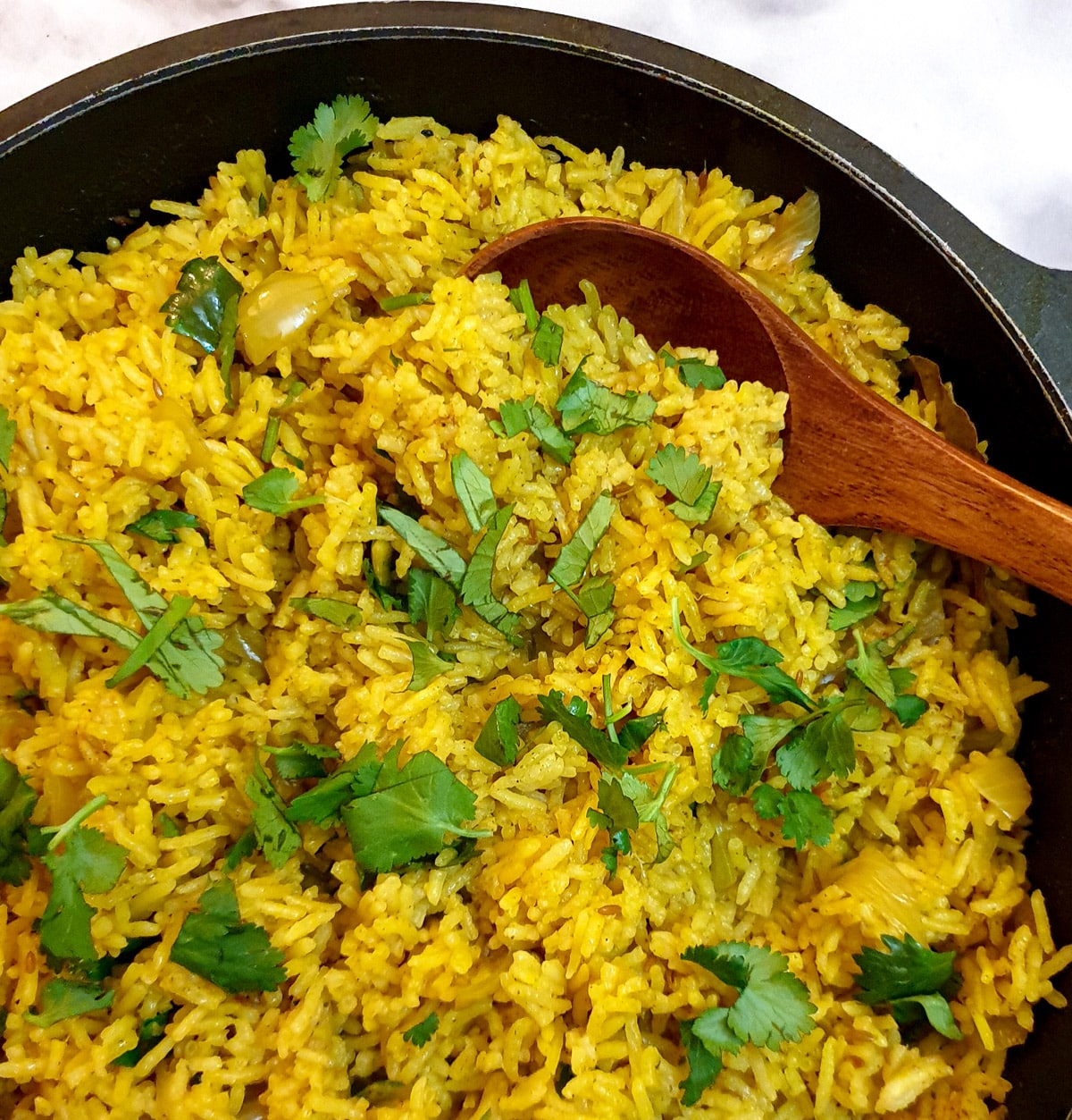 Close up of pilau rice in a black frying pan with a brown wooden serving spoon.