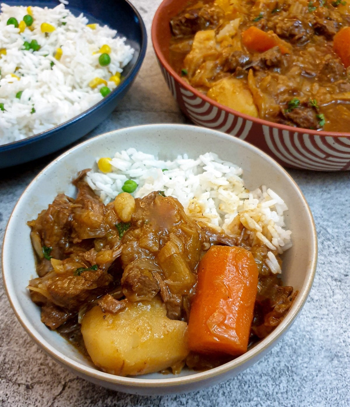A white bowl of beef bredie with rice and vegetables. In the background is a serving dish of stew and a dish of rice.