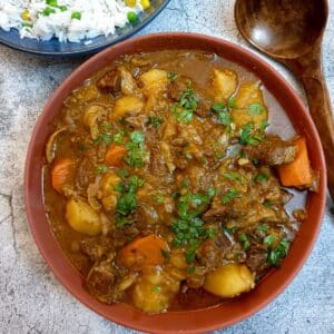 Overhead shot of a brown serving dish of South African beef stew with a dish of white rice and a brown wooden serving spoon.