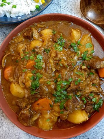 Overhead shot of a brown serving dish of South African beef stew with a dish of white rice and a brown wooden serving spoon.