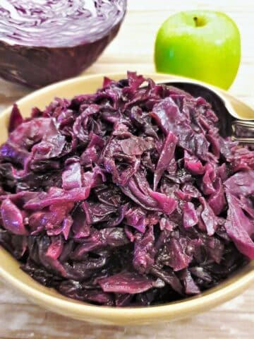 A dish of spicy braised red cabbage with an apple and raw red cabbage in the background.