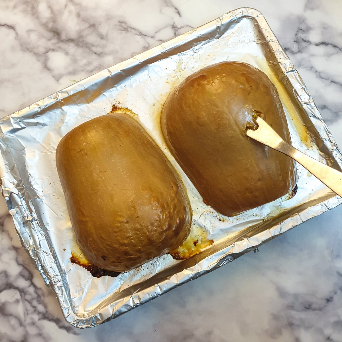 Two upside down butternuts on a baking tray, one of them being prodded with a long fork to show it is cooked.