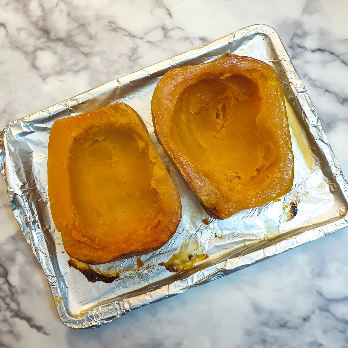 Two halved butternuts on a baking tray with the inside scooped out to leave only a half inch layer of butternut.