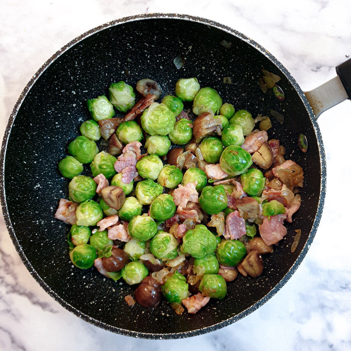 Brussels sprouts with bacon, onions and chestnuts in a black saucepan.