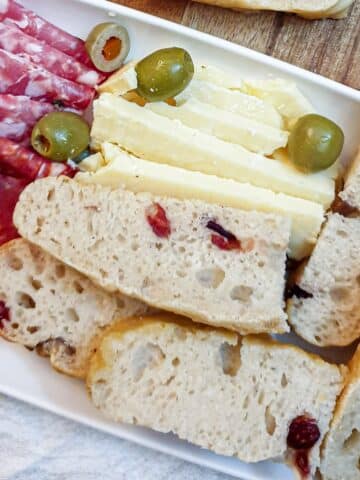 Slices of chestnut and cranberry focaccia of a long white plate with salami, cheese and olives.